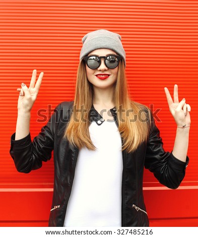 Fashion stylish cool girl having fun wearing a rock black leather jacket and sunglasses with hat over red background