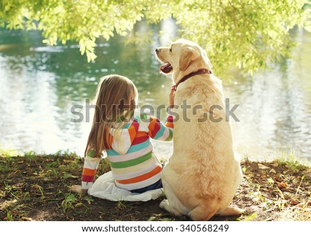Little child with Labrador retriever dog sitting in sunny summer park near water
