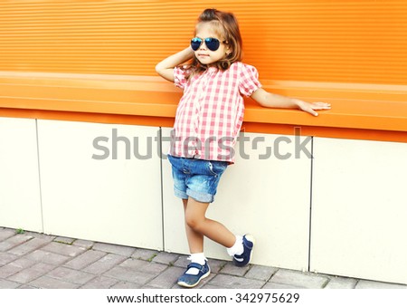Fashion little girl child wearing a sunglasses and checkered shirt over orange background in city