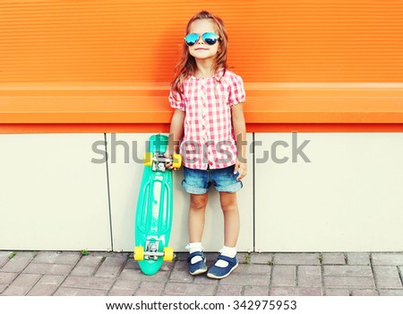 Stylish little girl child with skateboard wearing a sunglasses and checkered shirt over orange background