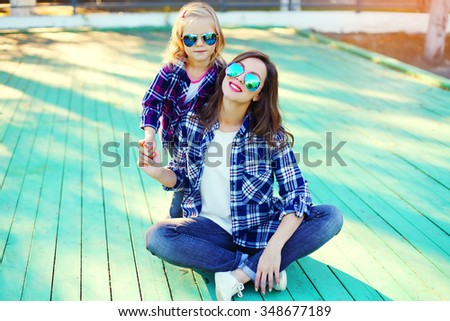 Fashion young mother and child daughter wearing a sunglasses and checkered shirt sitting in city