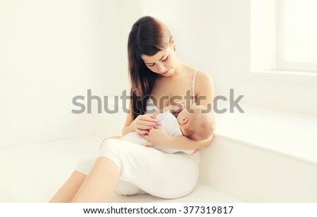 Young mother feeding breast her baby at home in white room near window