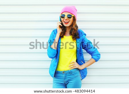 Fashion happy cool smiling girl talking on smartphone in colorful clothes over white background wearing pink hat yellow sunglasses and blue jacket