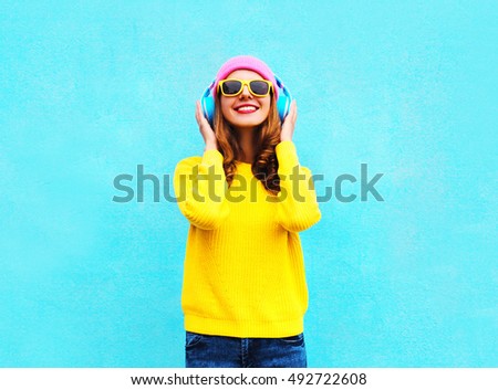 Pretty fashion cool smiling girl listening to music in headphones wearing a colorful pink hat, yellow sunglasses and sweater over blue background