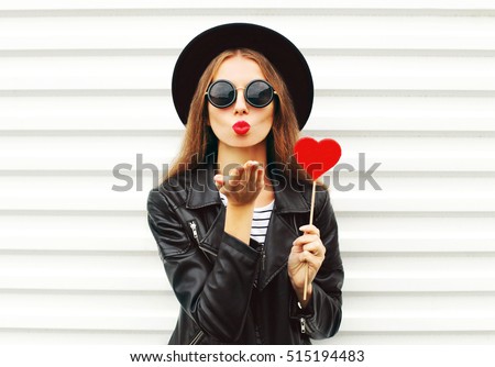 Fashion pretty sweet young woman with red lips sends air kiss with lollipop heart wearing black hat leather jacket over white background