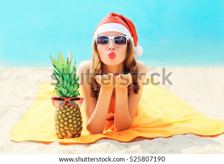 Christmas portrait pretty young woman in red santa hat with pineapple sends air kiss lying on beach over blue sea background