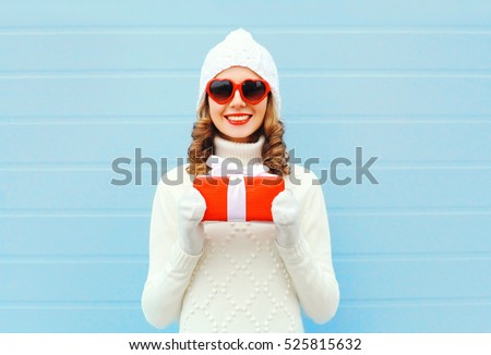 Christmas happy smiling young woman with gift box wearing a knitted hat sweater sunglasses over blue background