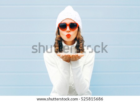 Happy woman blowing red lips sends air kiss wearing a heart shape sunglasses, knitted hat, sweater over blue background
