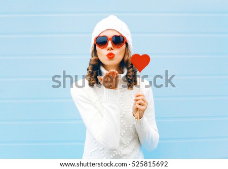 Portrait pretty woman blowing red lips sends air kiss holds lollipop heart wearing a heart shape sunglasses, knitted hat, sweater over blue background