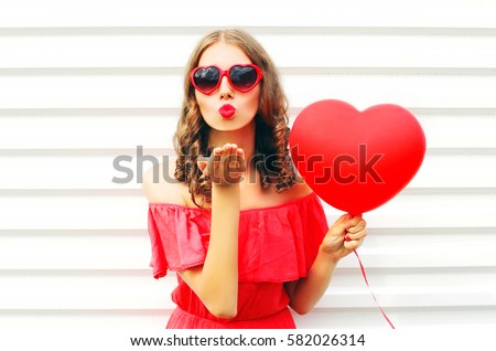 Portrait pretty woman in red dress sends air kiss with balloon heart shape over white background