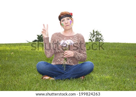 Mature woman dressed as a sixties flower child sitting in the grass holding daisies and flashing a peace sign
