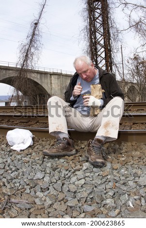 Homeless man sitting on edge of railroad tracks eating from a tin can holding a wine bottle in a paper bag