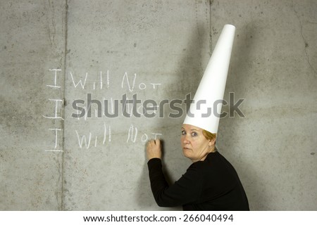 Woman wearing a dunce cap writing I WILL NOT on a concrete wall