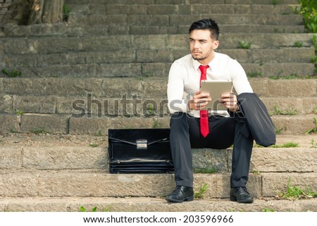 Young and successful worried business man sitting on the stairs in the park holding a tablet