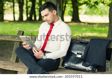 Young and successful worried business man sitting on the park bench holding a tablet