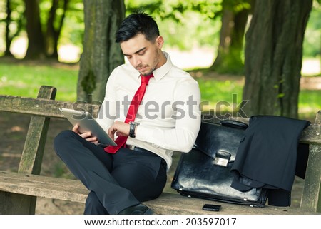 Young and successful business man sitting on the park bench holding a tablet and checking time
