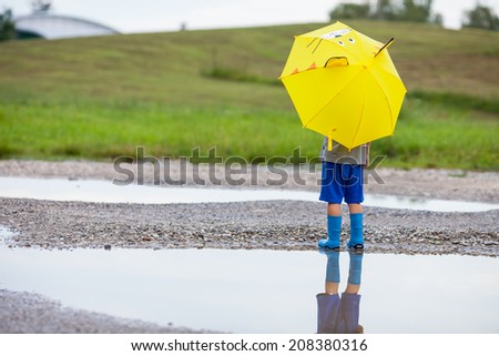 Cute little boy in the puddle with blue boots and yellow cat umbrella after the summer rain