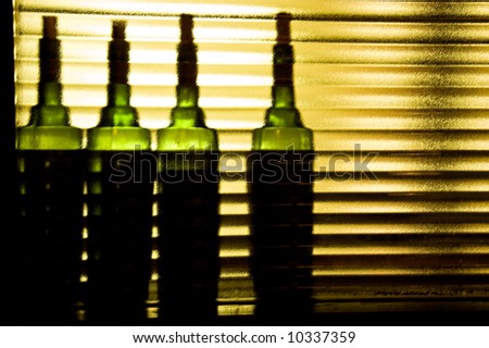 Four green bottles sitting behind a rippled gold-colored glass panel, lit by light coming through a tinted window.