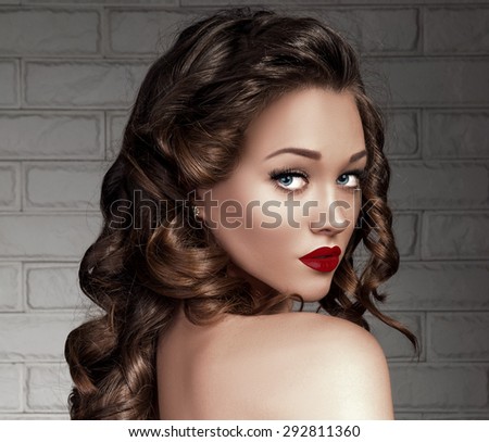 Beautiful Seductive Young Woman Model with Long Volume Curly Hair, Bronze Skin, Red Matte Sexy Lips, Blue Eyes and False Lashes. Trendy Evening Make up. Curls Hairstyle. Nude Shoulder.