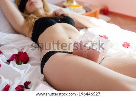 light pink or cream present & seductive blond beautiful young woman sexy romantic blindfolded girl in black lingerie relaxing lying on white bed with flower petals