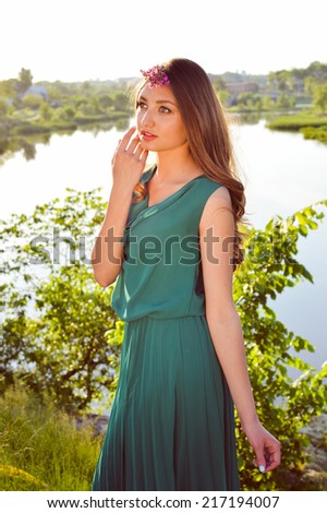portrait of young beautiful brunette lady in green dress & pink wreath of flowers having fun standing in grass against sun light & looking at outdoors copy space background