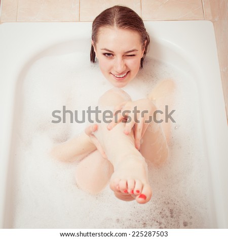 funny sniper in spa bath: portrait of beautiful blonde young sensual lady having fun happy smiling & enjoying aiming at camera with leg as a gun lying on foam water background