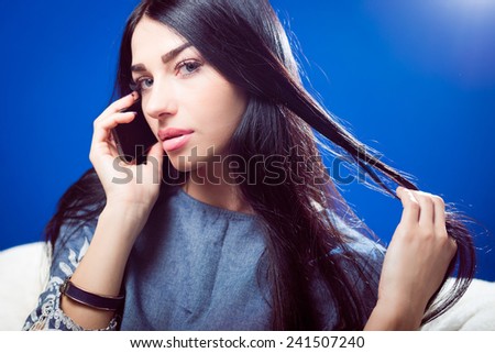 closeup portrait of pretty young lady talking on mobile phone and sensually looking at camera while touching luxury hair on blue copy space background