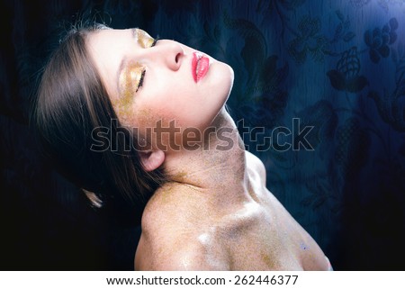 Close up image of covered in shining golden makeup sexy beautiful young lady with eyes closed relaxing on black printed pattern background