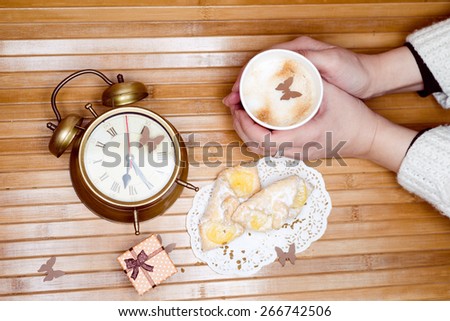 Image of hands holding cup of hot drink with pastries on plate, small gift box and alarm clock around