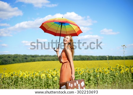 Portrait of beautiful young blond woman holding travel suitcase and rainbow umbrella on the sunflower field outdoors background copy space
