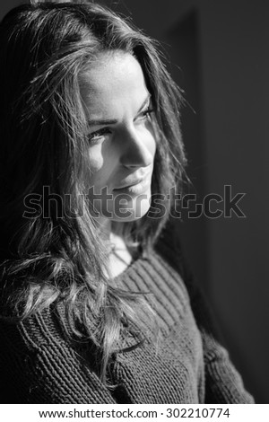 Beautiful young woman in woolen sweater enjoying sunshine and looking out the window. Black and white closeup portrait