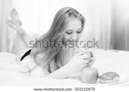 Black and white closeup portrait of beautiful sexy blonde young lady lying in bed smiling looking at plate with donut and apple on light window background