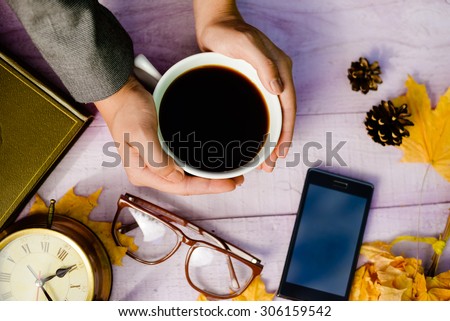 Top view close up on hands holding cup of coffee with mobile phone, glasses and alarm clock over copy space wooden table background