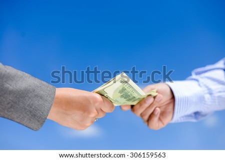 Picture of hands giving and receiving dollar bills. Side view in horizontal format over a blue sky sunny outdoors background.