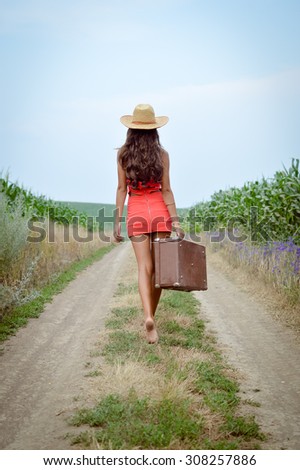 Female with gorgeous long hair, hat and short red dress walking away barefoot with retro suitcase along earth road in field on summer day outdoors background