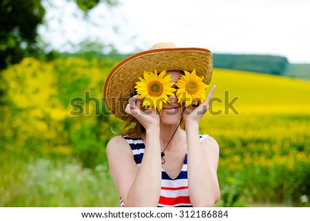 Young pretty lady wearing straw hat with two sunflowers instead glasses. Half length of smiling girl on countryside background.