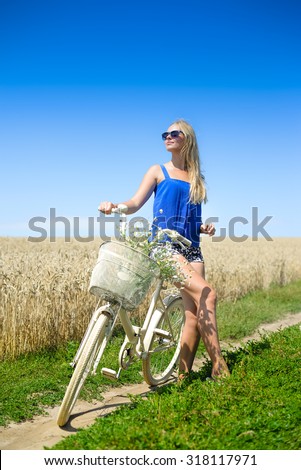 Picture of sexy blond girl on white bike with flower basket. Young girl in sunglasses relaxed on sunny summer countryside background.