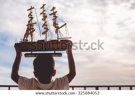 Picture of little boy holding beautiful ship model. Silhouette of happy kid on sunset sky outdoor background.