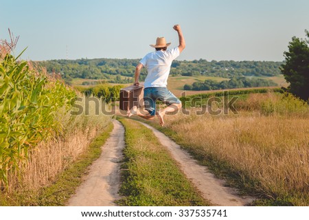Picture of man in straw hat holding old valize and jumping on country road. Back view of excited traveler on blurred sunny outdoor background.