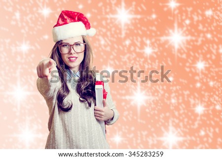 Picture of amazing beautiful young lady in red Santa Claus hat pointing finger and holding present box. Pretty girl in glasses happy smiling and looking at camera on abstract festive design background