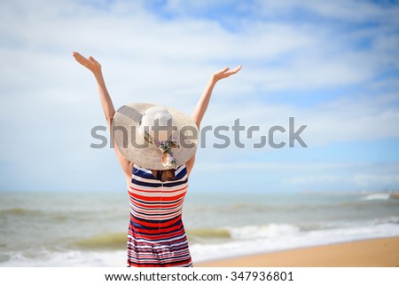 Back view photo of lady in straw hat and striped dress reaching her hands up and enjoying summer day on beautiful seaside