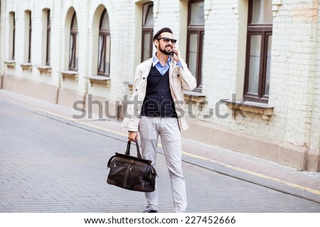 Young urban businessman on smart phone running in street talking on smartphone smiling wearing jacket and leather bag