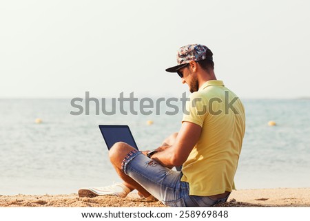 Young guy with a beard wearing glasses wearing a yellow T-shirt and shorts sits on a sandy ocean beach and working on laptop