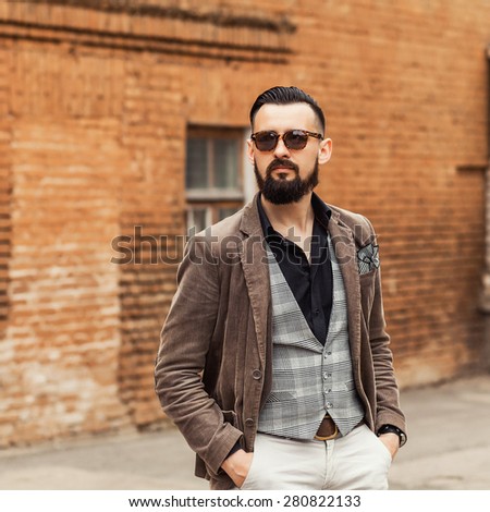 Young confident hipster man with beard in glasses posing on the street. Cute man wearing a vest, black shirt and light trousers. Man walks through the old town and looking forward