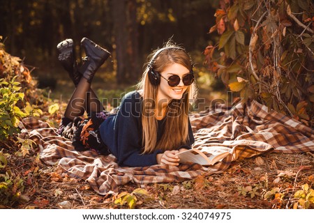 Young girl in headphones listening to music lying on a rug under the tree in the autumn forest. Girl in sunglasses dressed in a blue sweater and holding a book