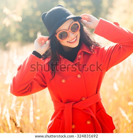 A beautiful young brunette in sunglasses smiling and standing in a field in autumn. Girl in a black hat and red coat