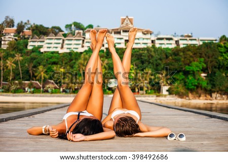 Two beautiful girls in swimsuits lying on the wooden pier upside down, against the background of a hotel on the beach