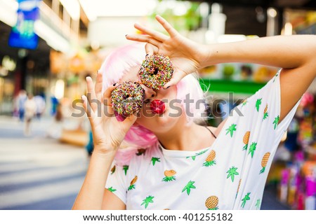 bright fashion funny girl in pink wig posing on background carousel at an amusement park. The hands holding the sweet donuts.