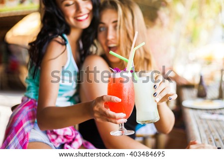 Two trendy cool hipster girls, friends, the blonde and the brunette drink cocktails at the cafe on the beach. Dressed in colorful shirts and shorts.