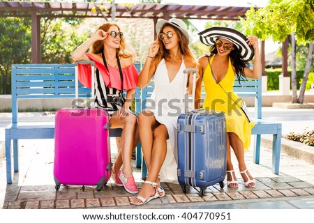 Three beautiful fashion girls sitting with their suitcases and wait in the terminal at the airport. Blonde, redhead, brunette, dressed in bright dresses and hats.